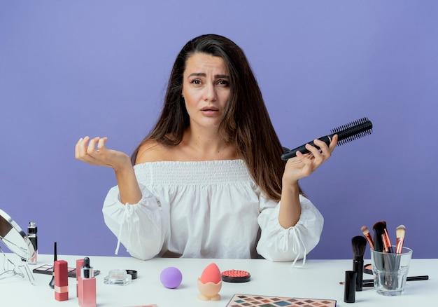 Woman looking frustrated while holding a hairbrush with makeup products on table