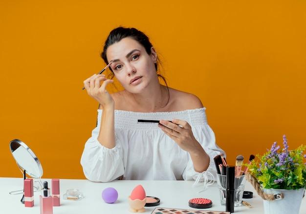 Woman holding makeup brush, sitting at cosmetic table with various beauty products, against an orange background